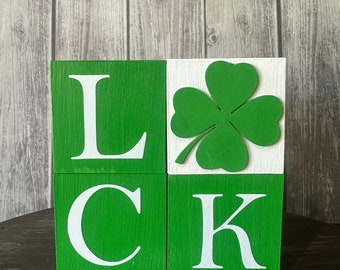 May include: Four wooden blocks spell out the word "LUCK" in white letters on a green background. The "C" block is in the lower left corner, the "K" block is in the lower right corner, and the "L" block is in the upper left corner. The "U" block is white with a green four-leaf clover cutout.