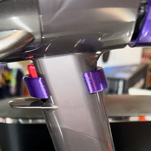 May include: Close-up of a cordless vacuum cleaner. The vacuum features a gray body with purple and red accents. The image highlights the handle, base, and attachment points. The vacuum is resting on a metallic surface.