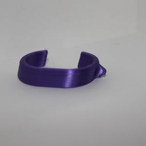 May include: A 3D-printed, purple, curved object with a small protrusion on one side. The object has a smooth, glossy finish and appears to be made of plastic. The design features two open ends, creating a U-shape.