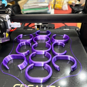 May include: A 3D printer bed with multiple purple, semi-circular objects. The objects are arranged in rows and columns. The printer has a black frame and the brand name "CREALITY" is visible on the bed.