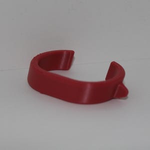 May include: A curved, red plastic object with a small protrusion on one end. The item has a smooth, matte finish and appears to be a clip or holder of some kind. The object is set against a plain white background.