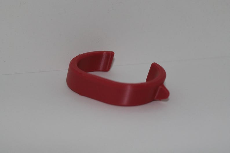 May include: A curved, red plastic object with a small protrusion on one end. The item has a smooth, matte finish and appears to be a clip or holder of some kind. The object is set against a plain white background.
