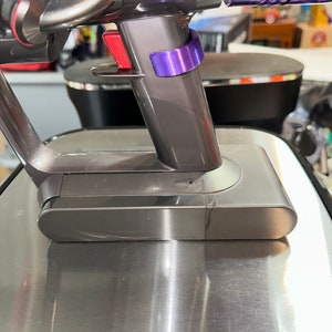 May include: Close-up of a cordless vacuum cleaner. The vacuum features a dark gray body with a red trigger and a purple accent. The base is a brushed metal finish. The image highlights the handle and base of the vacuum.