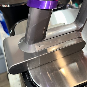 May include: Close-up of a cordless vacuum cleaner. The vacuum features a metallic gray body with a purple accent ring and a red button. The design is sleek and modern, suggesting a focus on efficiency and ease of use.