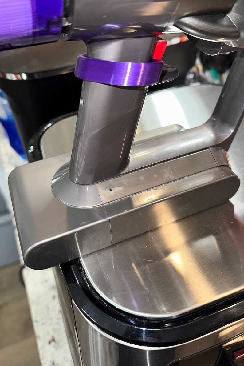 May include: Close-up of a cordless vacuum cleaner. The vacuum features a metallic gray body with a purple accent ring and a red button. The design is sleek and modern, suggesting a focus on efficiency and ease of use.