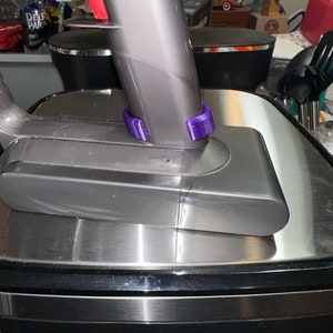 May include: A cordless vacuum cleaner with a gray body and a purple accessory. The vacuum is resting on a stainless steel surface. The image is a close-up of the vacuum's handle and base.