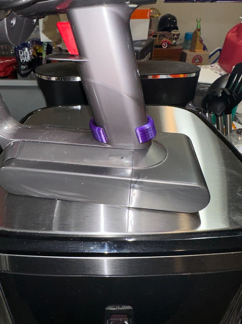 May include: A cordless vacuum cleaner with a gray body and a purple accessory. The vacuum is resting on a stainless steel surface. The image is a close-up of the vacuum's handle and base.