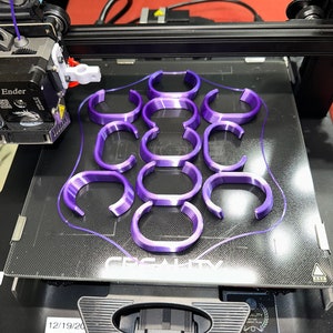 May include: A 3D printer in operation, printing multiple purple, curved objects. The objects are arranged in a grid pattern on the printer bed. The printer is a black and silver Ender model.