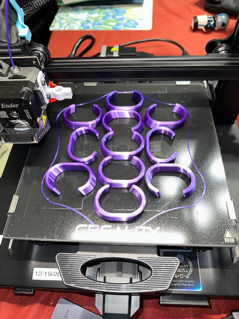 May include: A 3D printer in operation, printing multiple purple, curved objects. The objects are arranged in a grid pattern on the printer bed. The printer is a black and silver Ender model.