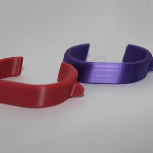 May include: Two curved, open-ended plastic rings. One is a solid red, the other is a gradient of purple. The rings have a small, raised tab on one side. The objects are on a white surface.