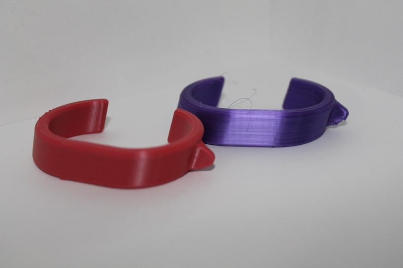 May include: Two curved, open-ended plastic rings. One is a solid red, the other is a gradient of purple. The rings have a small, raised tab on one side. The objects are on a white surface.