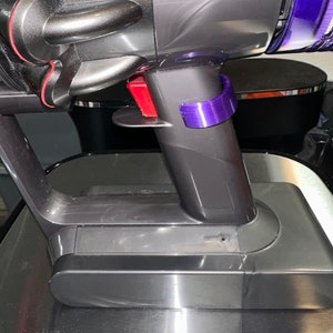 May include: Close-up of a gray and purple vacuum cleaner. The vacuum has a red button and a purple clip. The vacuum is on a metallic surface.