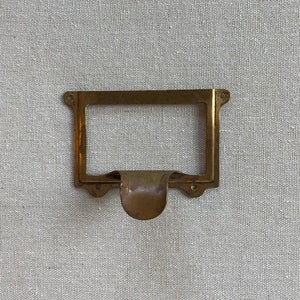 Puede incluir: Un tirador de cajón de latón con un marco rectangular y una manija curva. El tirador tiene un aspecto vintage y sería una gran adición a cualquier hogar.