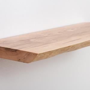 Puede incluir: Una estantería de madera natural con un borde vivo y un color marrón claro. La estantería está hecha de una sola pieza de madera y tiene un aspecto rústico.