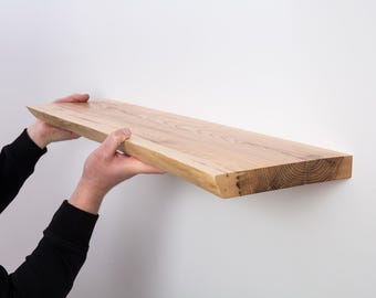 Può includere: Una persona sta tenendo in alto un lungo ripiano in legno naturale con un bordo vivo. Il ripiano è di colore marrone chiaro e ha una texture ruvida e non rifinita.