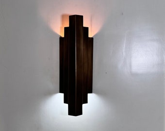 May include: A bronze wall sconce with a geometric design. The sconce is lit and casting a warm glow on the white wall.