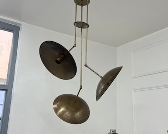 May include: A brass-toned, mid-century modern chandelier with three disc-shaped shades suspended from a central fixture. The shades are angled, and the fixture is mounted on a white ceiling. A spotlight on a tripod is in the background.