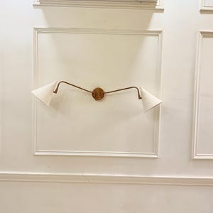 May include: A wall-mounted light fixture with a circular wooden base and two curved, bronze-colored arms. Each arm supports a white, triangular lampshade. The fixture is mounted on a white wall with decorative molding.