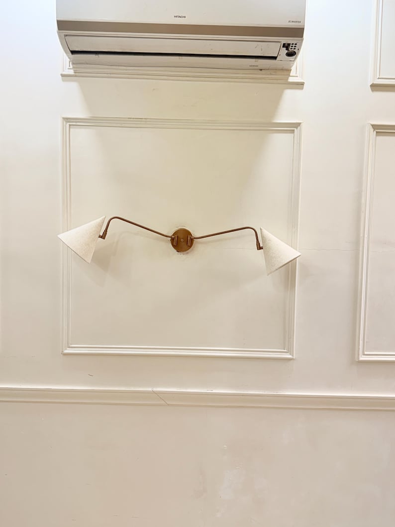 May include: A wall-mounted light fixture with a circular wooden base and two curved, bronze-colored arms. Each arm supports a white, triangular lampshade. The fixture is mounted on a white wall with decorative molding.