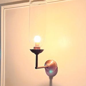 May include: A wall-mounted sconce with a clear glass shade and a warm, glowing light bulb. The sconce has a dark bronze-colored decorative base and arm, and a copper-colored wall plate. The fixture is mounted on a light-colored wall.