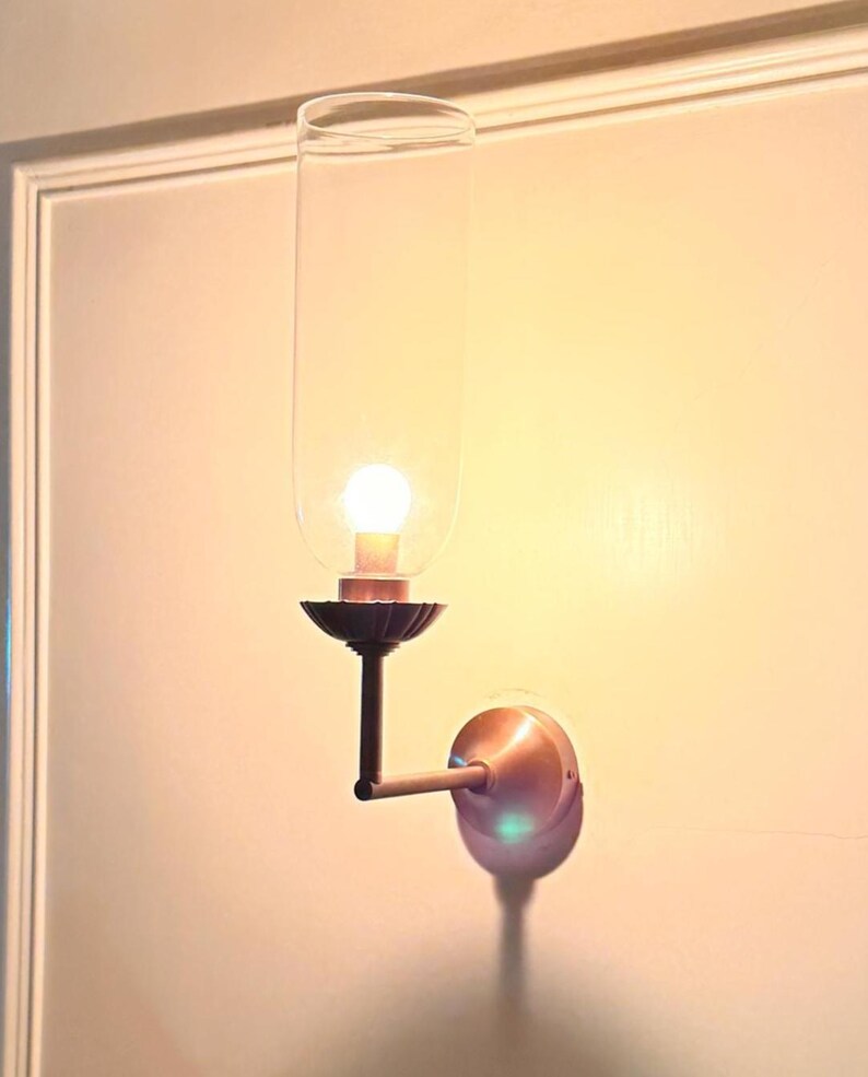 May include: A wall-mounted sconce with a clear glass shade and a warm, glowing light bulb. The sconce has a dark bronze-colored decorative base and arm, and a copper-colored wall plate. The fixture is mounted on a light-colored wall.