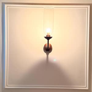 May include: A wall-mounted sconce with a clear glass shade and a warm, glowing light bulb. The sconce has a dark metal base and is mounted on a white wall with a framed border. The light casts a shadow below.