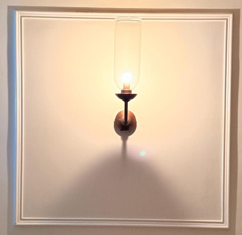 May include: A wall-mounted sconce with a clear glass shade and a warm, glowing light bulb. The sconce has a dark metal base and is mounted on a white wall with a framed border. The light casts a shadow below.