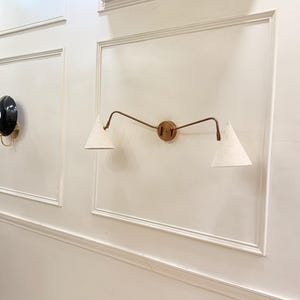May include: A wall-mounted light fixture with a circular brass base and two curved arms. Each arm supports a white, cone-shaped lampshade. The fixture is mounted on a white wall with decorative molding. A black and gold sconce is visible on the left.