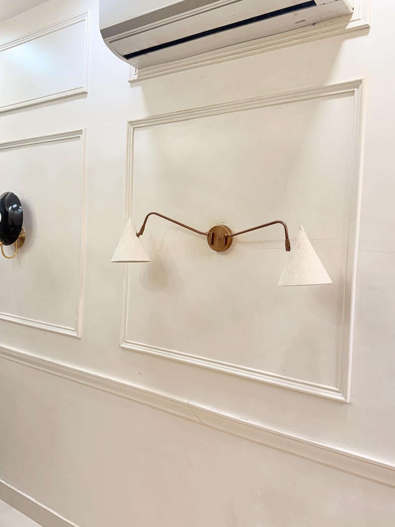 May include: A wall-mounted light fixture with a circular brass base and two curved arms. Each arm supports a white, cone-shaped lampshade. The fixture is mounted on a white wall with decorative molding. A black and gold sconce is visible on the left.
