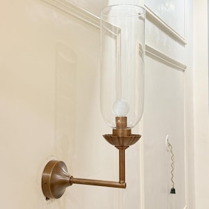 May include: A wall-mounted sconce with a clear glass shade and a bronze-colored metal base. The sconce features a decorative, fluted base and a long, straight arm. A light bulb is visible inside the glass shade. The sconce is mounted on a cream-colored wall.