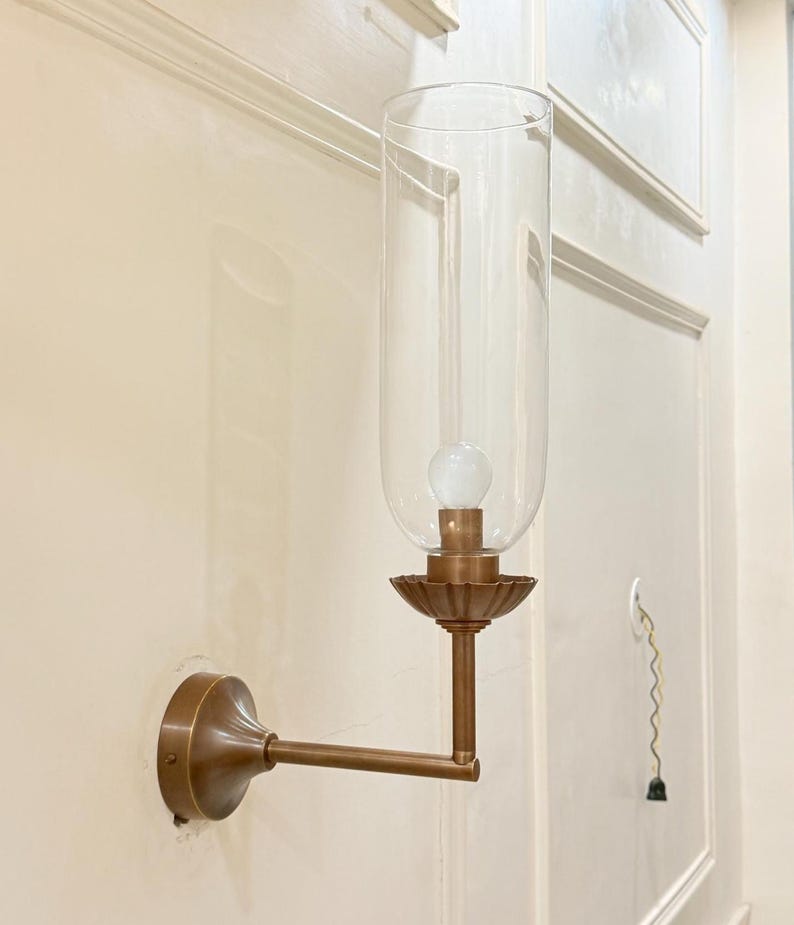 May include: A wall-mounted sconce with a clear glass shade and a bronze-colored metal base. The sconce features a decorative, fluted base and a long, straight arm. A light bulb is visible inside the glass shade. The sconce is mounted on a cream-colored wall.