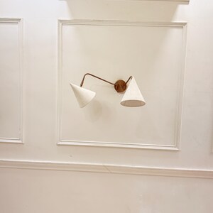 May include: A wall-mounted light fixture with two cone-shaped shades. The fixture has a bronze-colored arm and a circular base. The shades are a light beige color, and the fixture is mounted on a white wall.