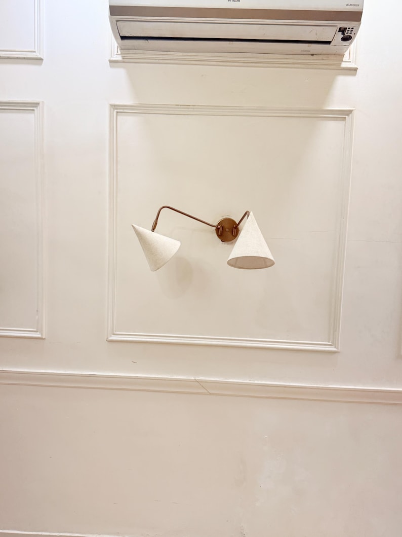 May include: A wall-mounted light fixture with two cone-shaped shades. The fixture has a bronze-colored arm and a circular base. The shades are a light beige color, and the fixture is mounted on a white wall.