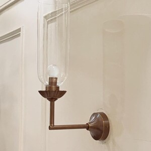 May include: A wall-mounted light fixture with a clear glass shade and a bronze-colored metal frame. The fixture has a bulb inside the glass shade and is mounted on a white wall. The fixture's arm extends horizontally.