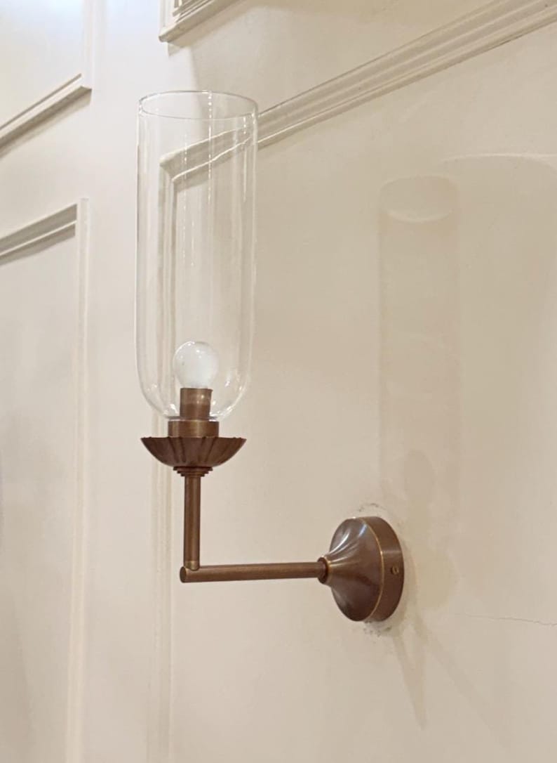May include: A wall-mounted light fixture with a clear glass shade and a bronze-colored metal frame. The fixture has a bulb inside the glass shade and is mounted on a white wall. The fixture's arm extends horizontally.