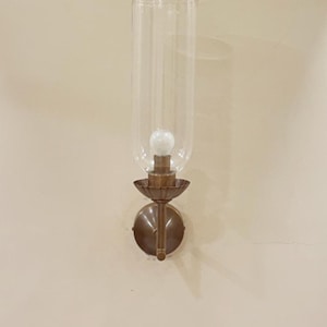 May include: A wall-mounted sconce with a clear glass shade and a bronze-colored metal base. The sconce features a bulb inside the glass shade, and a decorative, circular backplate. The fixture is mounted on a light beige wall.