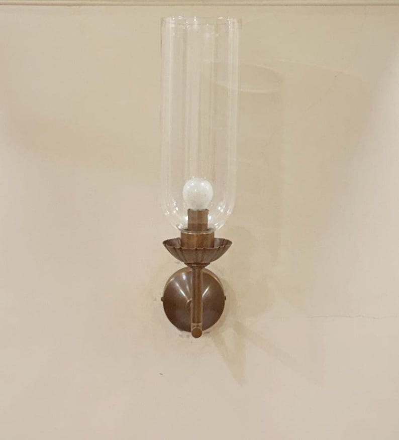 May include: A wall-mounted sconce with a clear glass shade and a bronze-colored metal base. The sconce features a bulb inside the glass shade, and a decorative, circular backplate. The fixture is mounted on a light beige wall.