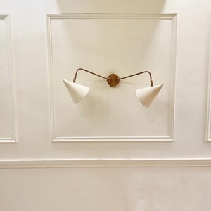 May include: A wall-mounted light fixture with a bronze finish and two cone-shaped, off-white fabric shades. The fixture is mounted on a white wall with decorative molding, creating a minimalist and modern aesthetic.