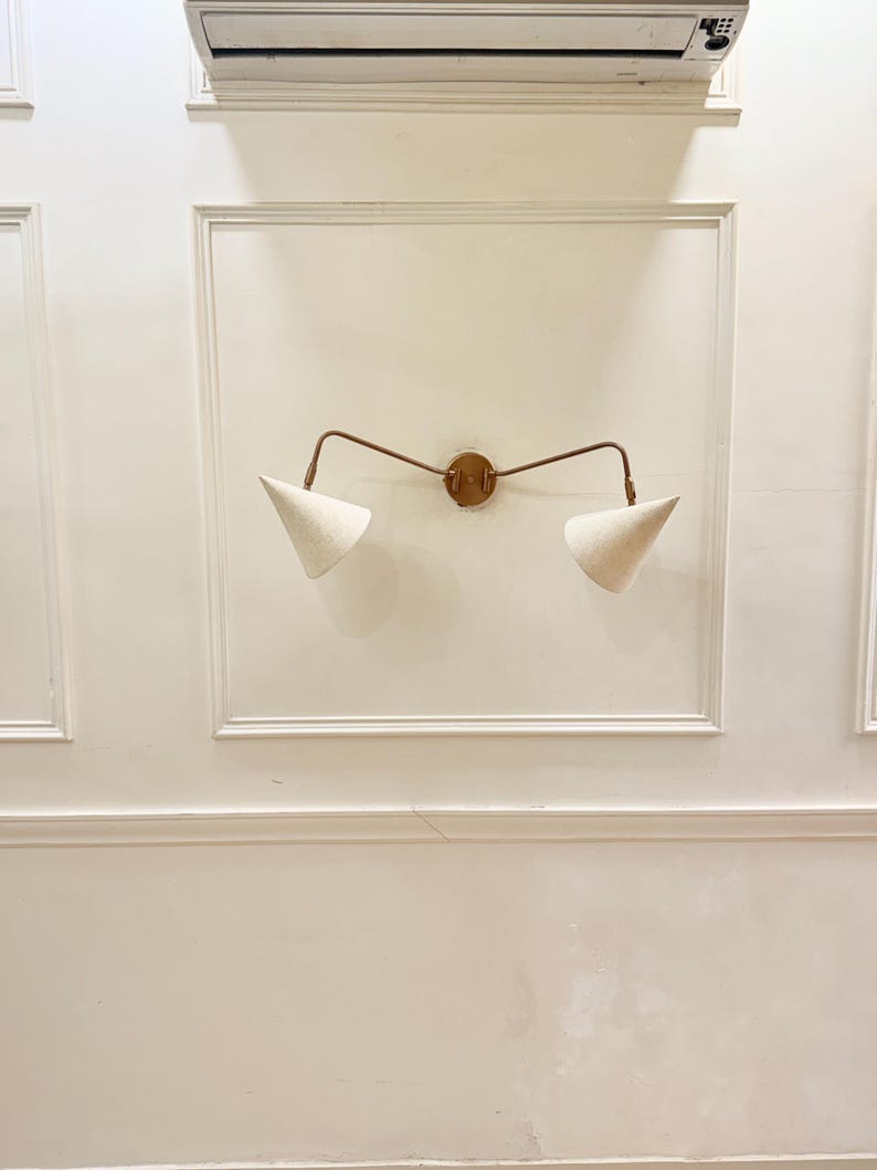 May include: A wall-mounted light fixture with a bronze finish and two cone-shaped, off-white fabric shades. The fixture is mounted on a white wall with decorative molding, creating a minimalist and modern aesthetic.