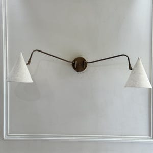 May include: A wall-mounted light fixture with two cone-shaped shades. The shades are a light beige color, and the fixture has a bronze finish. The fixture is mounted on a white wall.