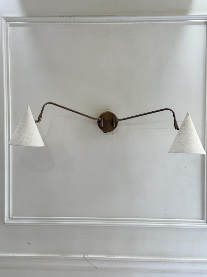 May include: A wall-mounted light fixture with two cone-shaped shades. The shades are a light beige color, and the fixture has a bronze finish. The fixture is mounted on a white wall.