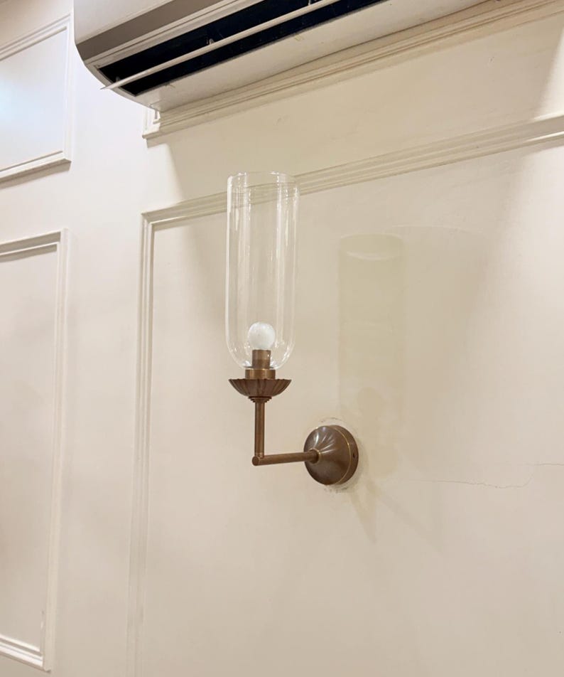 May include: A wall-mounted sconce with a clear glass shade and a bronze-colored metal base. The sconce features a bulb inside the glass shade and is mounted on a cream-colored wall.