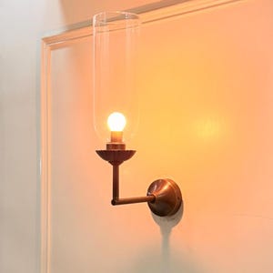 May include: A wall-mounted light fixture with a clear glass shade and a warm, glowing bulb. The fixture has a bronze-colored metal base and arm, mounted on a white wall. The design is reminiscent of vintage or antique lighting.