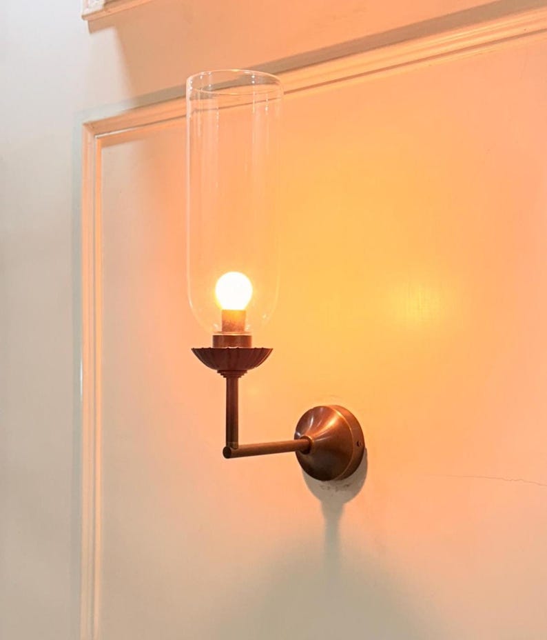 May include: A wall-mounted light fixture with a clear glass shade and a warm, glowing bulb. The fixture has a bronze-colored metal base and arm, mounted on a white wall. The design is reminiscent of vintage or antique lighting.
