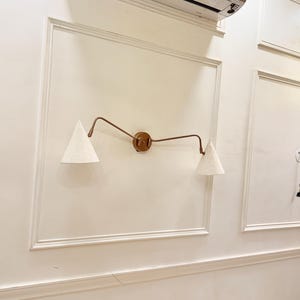 May include: A wall-mounted light fixture with two cone-shaped lampshades. The fixture has a bronze-colored frame and a circular base. The lampshades are white and textured, and the fixture is mounted on a cream-colored wall.