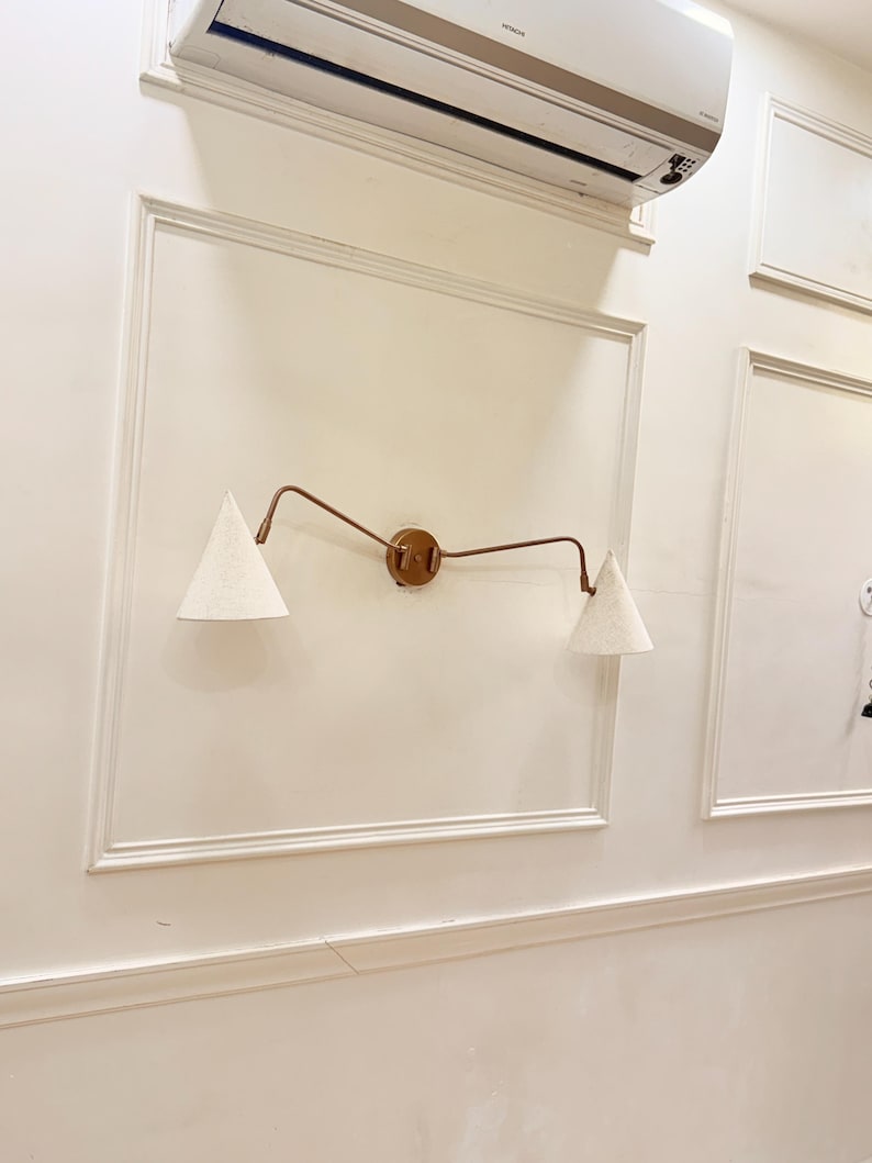 May include: A wall-mounted light fixture with two cone-shaped lampshades. The fixture has a bronze-colored frame and a circular base. The lampshades are white and textured, and the fixture is mounted on a cream-colored wall.
