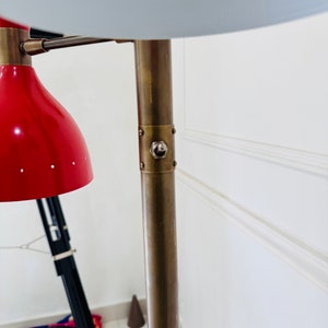 May include: A brass floor lamp with two shades. The top shade is white with holes and the bottom shade is red with holes. The lamp has a brass base and a brass pole.
