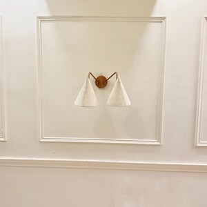 May include: A wall-mounted sconce with a bronze-colored frame and two conical, off-white fabric shades. The sconce is centered on a white wall with decorative molding.