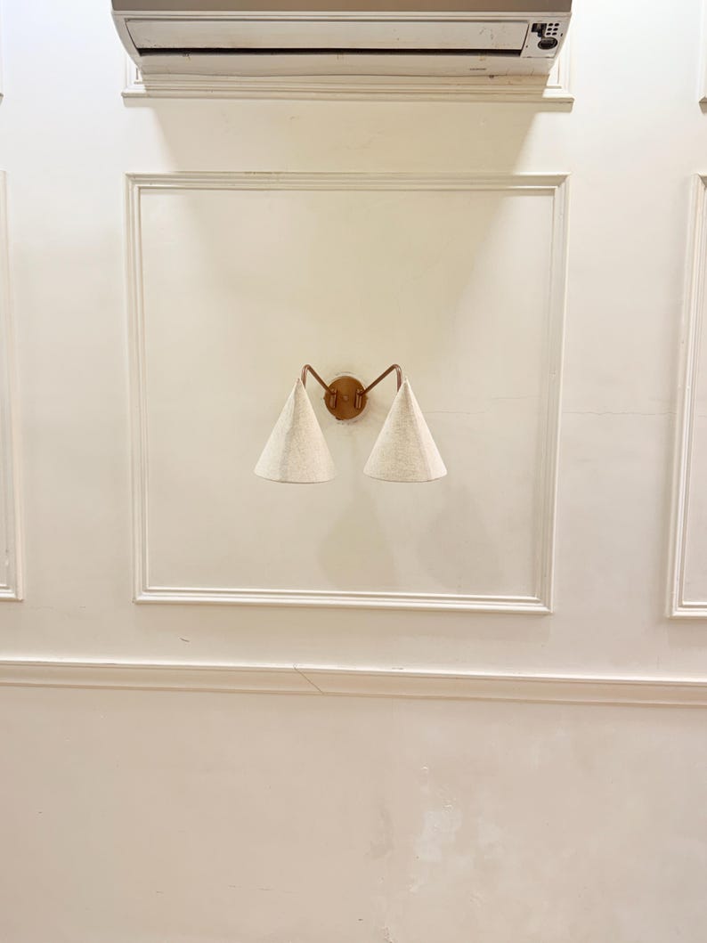 May include: A wall-mounted sconce with a bronze-colored frame and two conical, off-white fabric shades. The sconce is centered on a white wall with decorative molding.