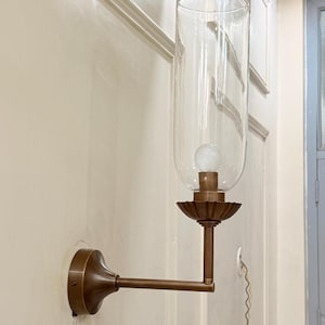 May include: A vintage-style wall sconce with a clear glass shade and a bronze-colored metal frame. The sconce features a decorative base and a long, straight arm extending from the wall. An exposed light bulb is visible inside the glass shade.